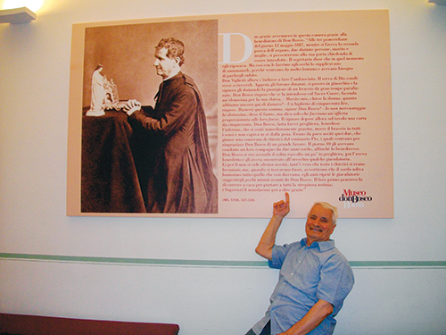 Photographie du père Cosimo montrant du doigt une fresque représentant Don Bosco à genoux et en prière